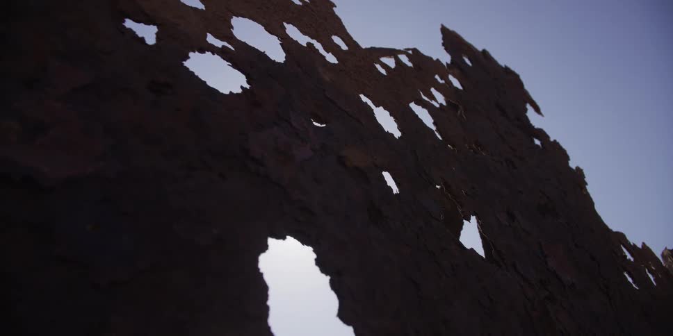 An abstract shot tracks along a heavily corroded and rusted metal sheet full of holes, silhouetted against a clear sky