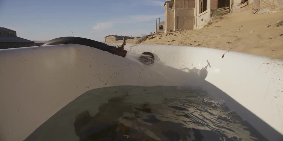 Water sprays from a rusty hose into an old bathtub sitting in the middle of a sandy, abandoned desert town