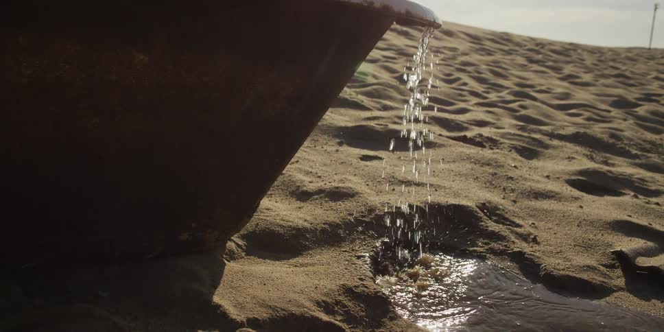 Water pours in slow motion from an old, discarded bathtub onto the golden sand of a barren desert