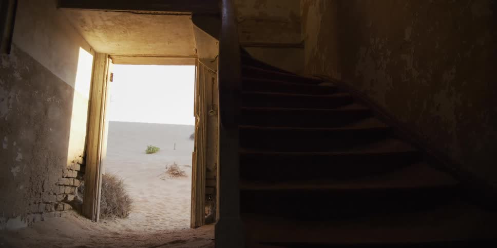 A slow pull-back shot reveals a dark, abandoned staircase inside a house being reclaimed by the desert sand