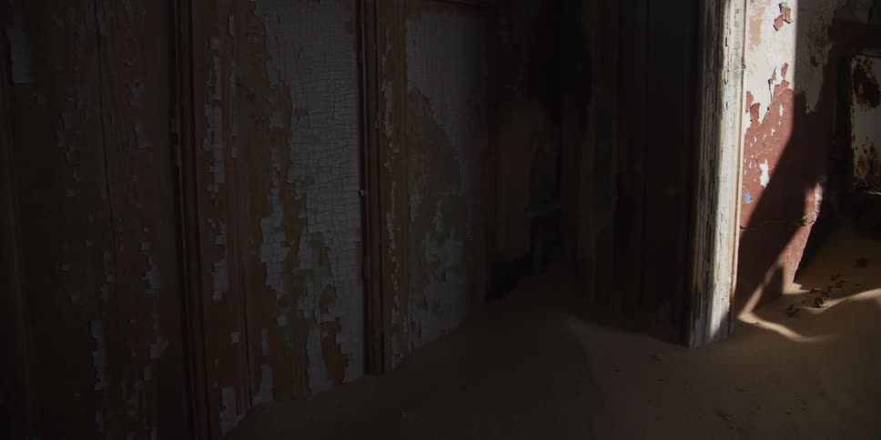 A slow dolly shot reveals the sand-filled interior of a decrepit, abandoned building with peeling paint on the walls