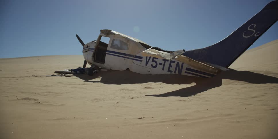 A wrecked propeller plane lies half-buried in the sand of a vast desert under a clear, sunny sky