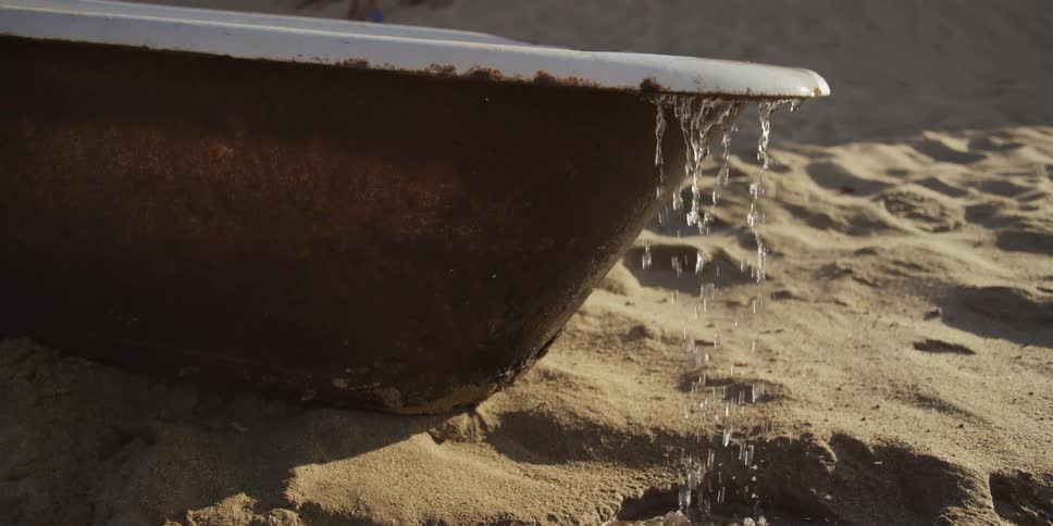 Water overflows in slow motion from an old rusty bathtub sitting on a sandy beach