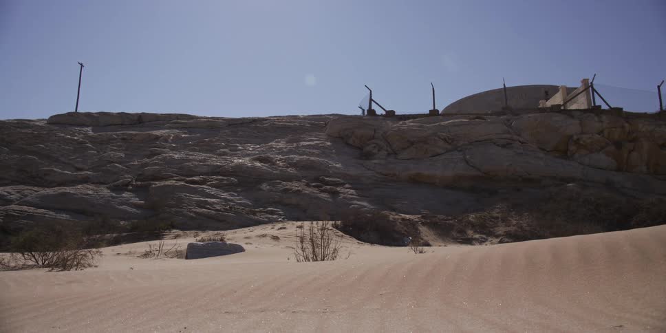 Still shot of a desolate, sun-bleached desert landscape with wind gently blowing sand across the foreground