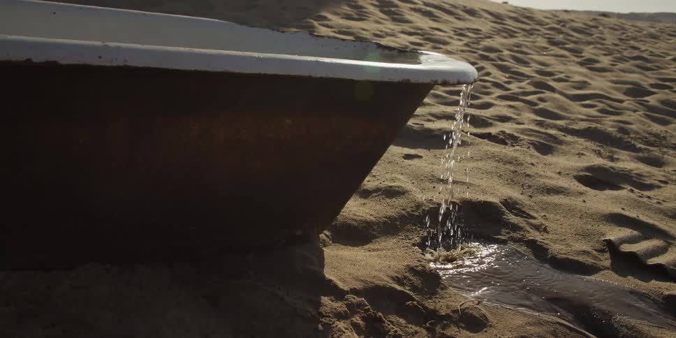 Water overflows from a rusty, abandoned bathtub onto the sand in slow motion