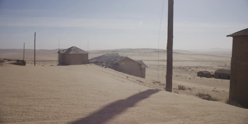 Desolate ghost town buildings are consumed by shifting desert sands under a harsh sun