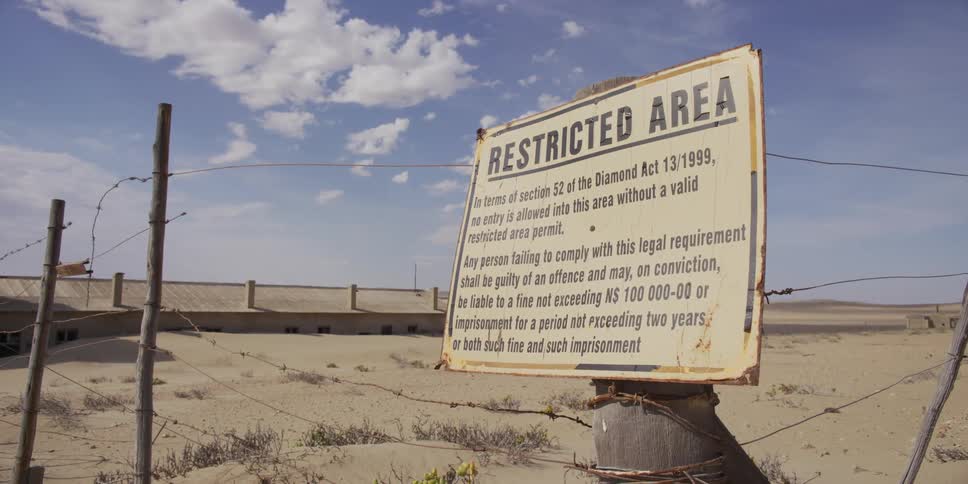 A weathered "Restricted Area" sign for a diamond mine stands on a barbed-wire fence in the desolate Namibian desert