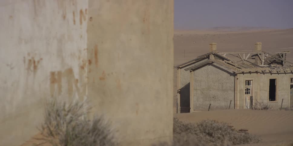 A rack focus shot reveals a dilapidated, abandoned house partially buried by sand in a desolate desert ghost town