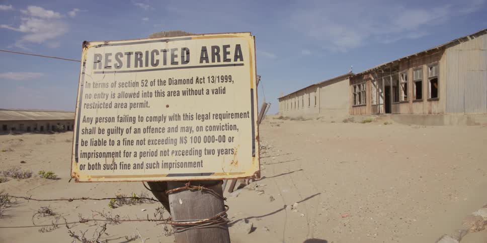 A slow zoom reveals a weathered 'Restricted Area' sign for a former diamond mine standing before derelict buildings in the Namibian desert