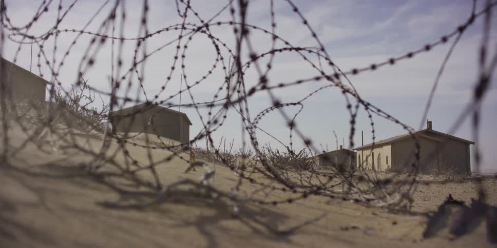 A low-angle view shows a coil of barbed wire on a sandy dune as wind blows past an abandoned desert outpost