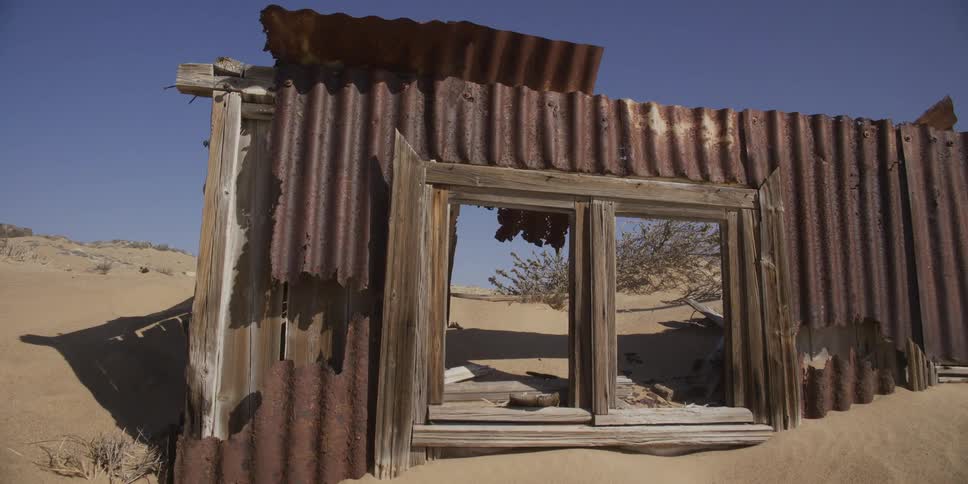 Slow pullback shot reveals a rusted, dilapidated shack with a wooden window frame being slowly buried by the desert sand