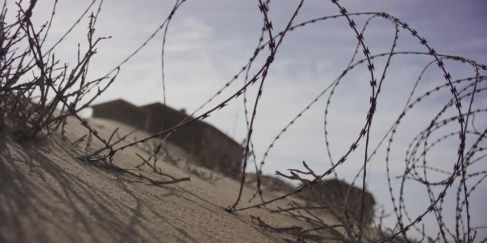 A low-angle close-up captures wind blowing sand over a dune past coiled barbed wire, with a desolate building in the background