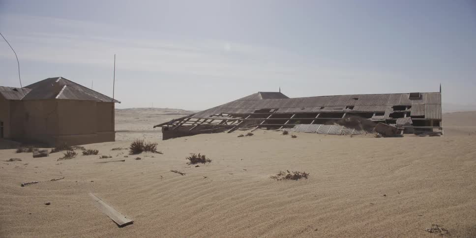 Wind blows sand over dilapidated, abandoned buildings being reclaimed by the vast, empty desert