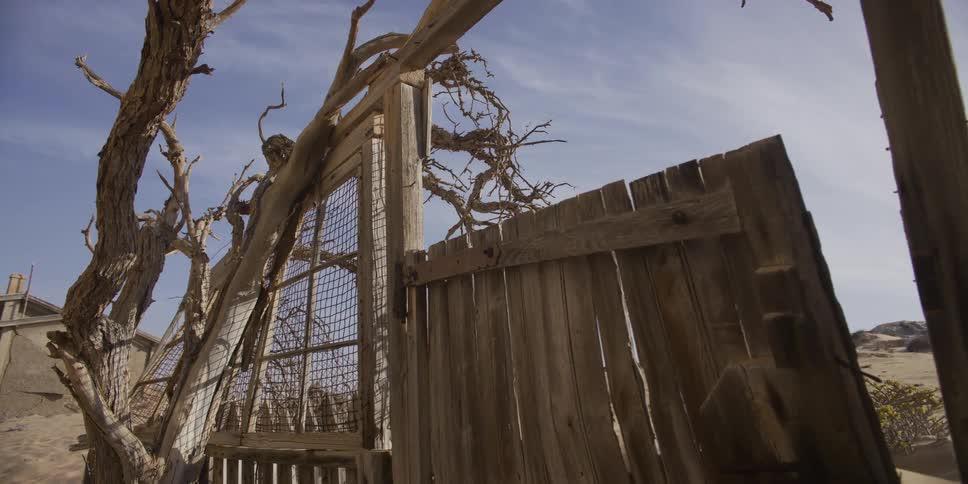 A slow pull-back shot reveals a desolate, abandoned wooden shack partially buried in the shifting desert sand