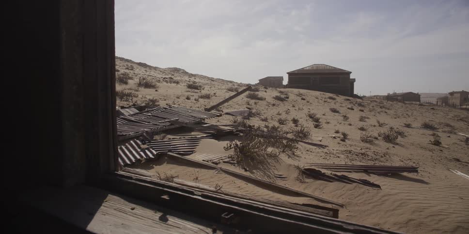 A slow pull-back through a dilapidated window frame reveals the sandy, desolate landscape of an abandoned ghost town