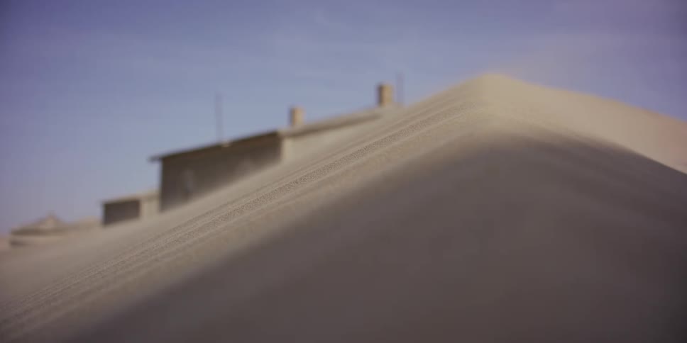 A close up shot shows sand blowing in the wind over a dune, slowly burying an abandoned building in the desert