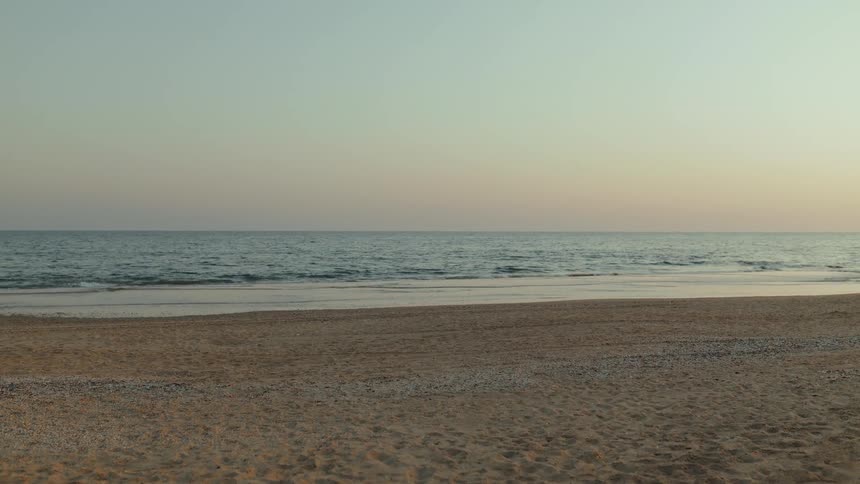 A peaceful, wide shot of a sandy beach with calm ocean waves lapping the shore at dusk