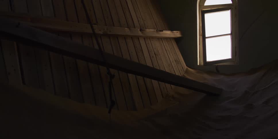 The dark interior of an abandoned wooden house is slowly being buried by sand, with a single window illuminating the eerie scene