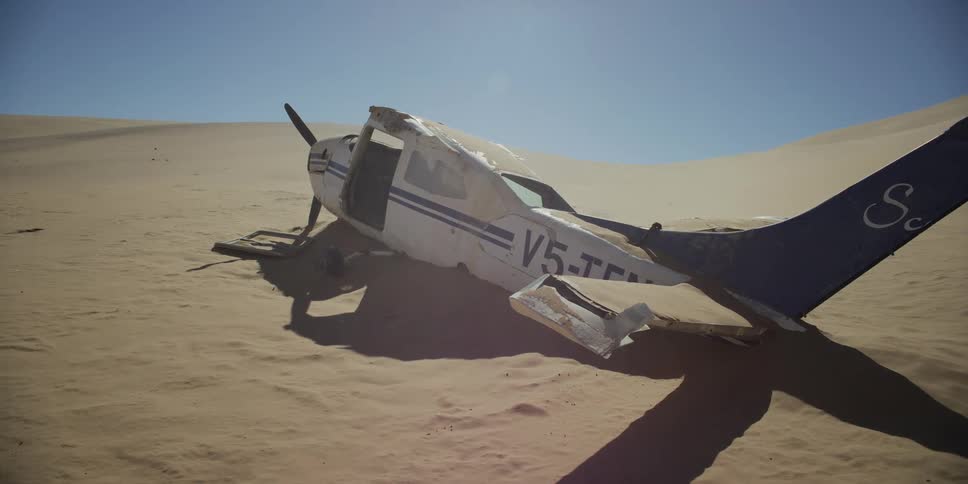 A crashed and abandoned propeller plane lies half-buried in a vast, sun-drenched desert as wind blows sand across the wreckage