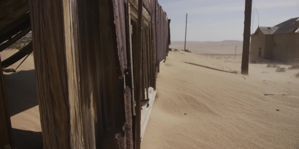 Close-up of a weathered, rusty building in an abandoned desert ghost town, with a slow pan revealing the sand-swept landscape