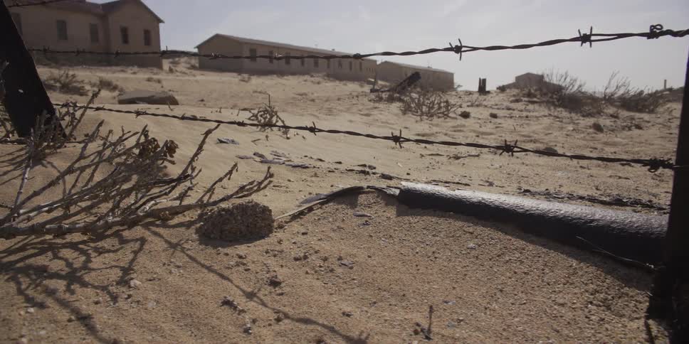 A slow dolly shot past a rusted barbed wire fence in the desert with abandoned buildings in the background