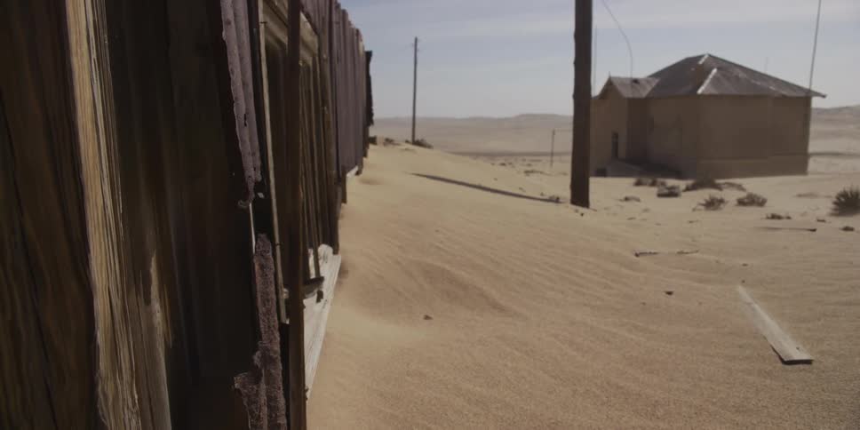 A slow pan reveals abandoned, dilapidated buildings in a desert ghost town being slowly consumed by shifting sands