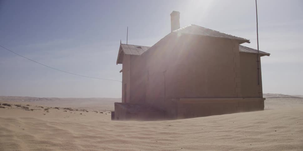 Wind blows sand across a desolate desert landscape, surrounding an abandoned house as the sun shines brightly
