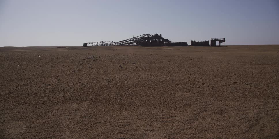 A wide, static shot shows the decaying skeleton of an abandoned industrial complex in a vast, empty desert