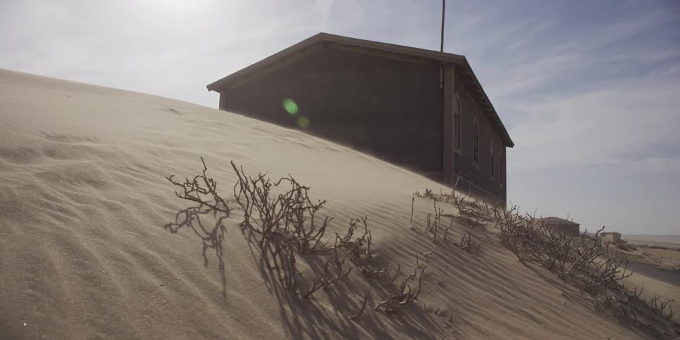 A slow dolly shot reveals a windswept sand dune slowly burying an abandoned desert building in a ghost town