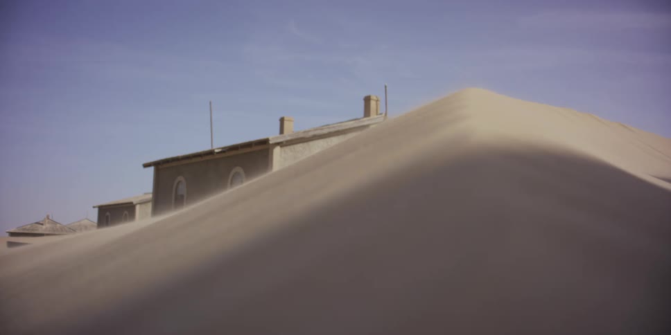 An abandoned desert home is slowly consumed by a massive, wind-swept sand dune, with a rack focus to the sandy crest