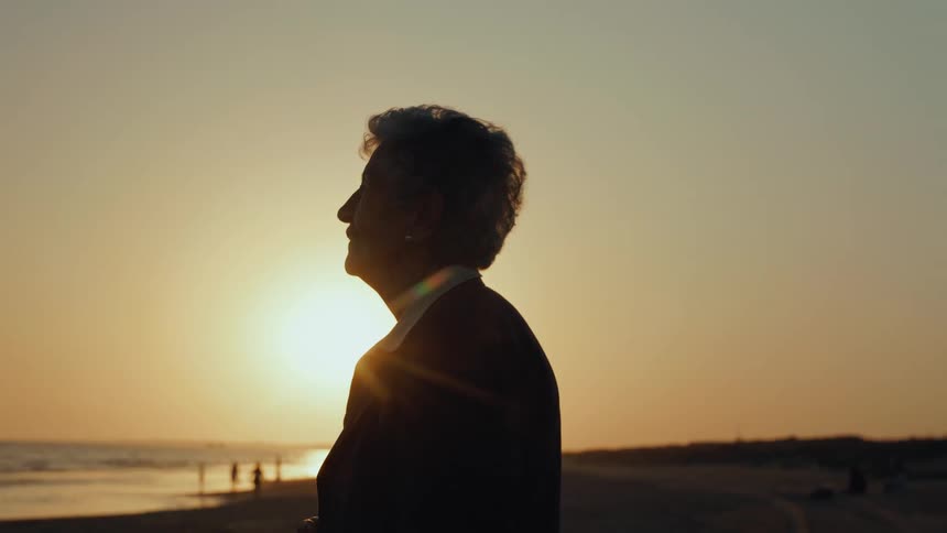 A silhouetted senior woman peacefully watches the golden sunset over the ocean from the beach