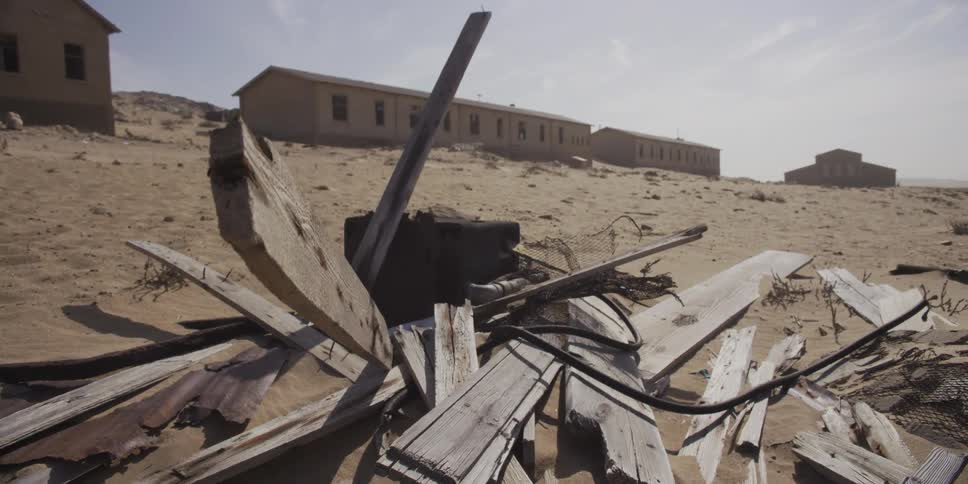 A slow dolly shot pulls back from weathered wooden debris to reveal an abandoned ghost town being reclaimed by the desert sand