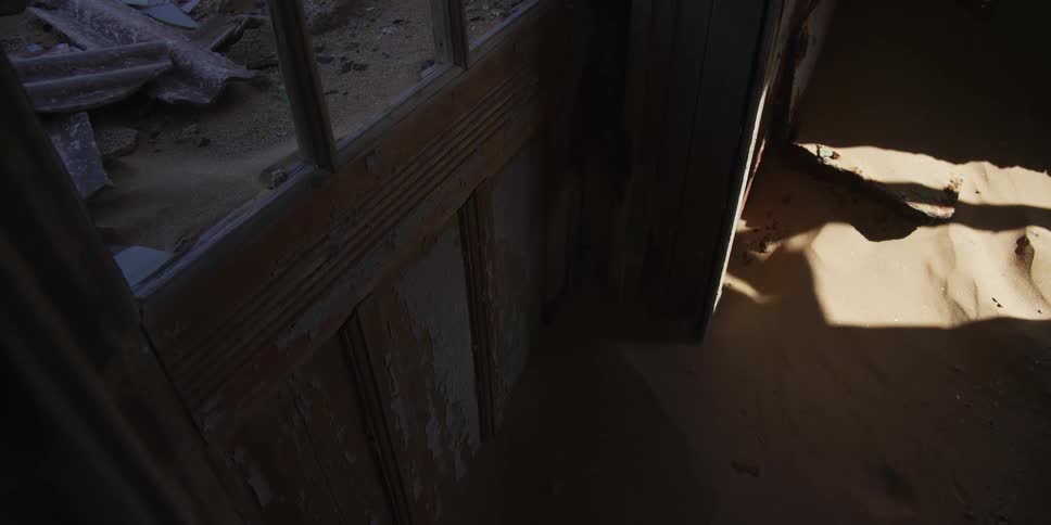 A slow reveal from a broken window shows an abandoned desert house being reclaimed by nature as sand fills the decaying room