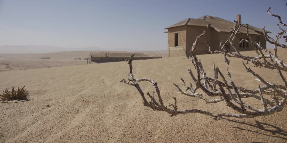 Slow dolly shot past a dead bush reveals an abandoned building being reclaimed by the vast desert