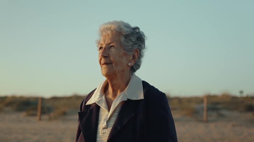 A serene elderly woman stands on a sandy beach, smiling thoughtfully as she looks towards the warm golden hour light