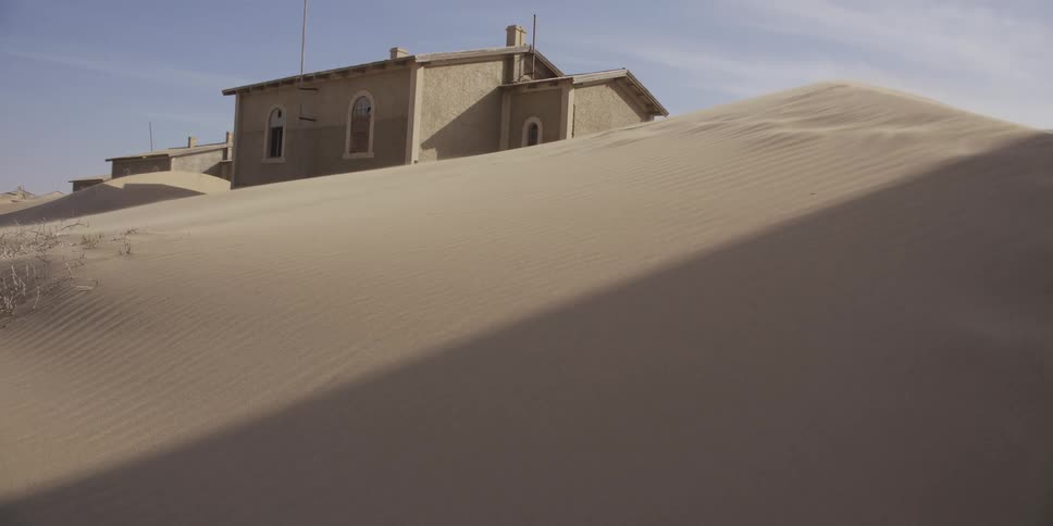 Sunlight and shadows move across a vast sand dune that is slowly burying an abandoned building in the ghost town of Kolmanskop, Namibia