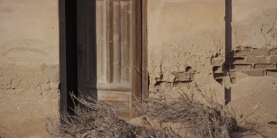 A weathered wooden door on an abandoned adobe building is partially buried by sand and tumbleweeds in a desert ghost town