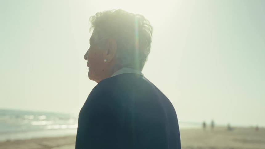 A pensive elderly woman with grey hair walks slowly towards the tranquil ocean on a sunny day