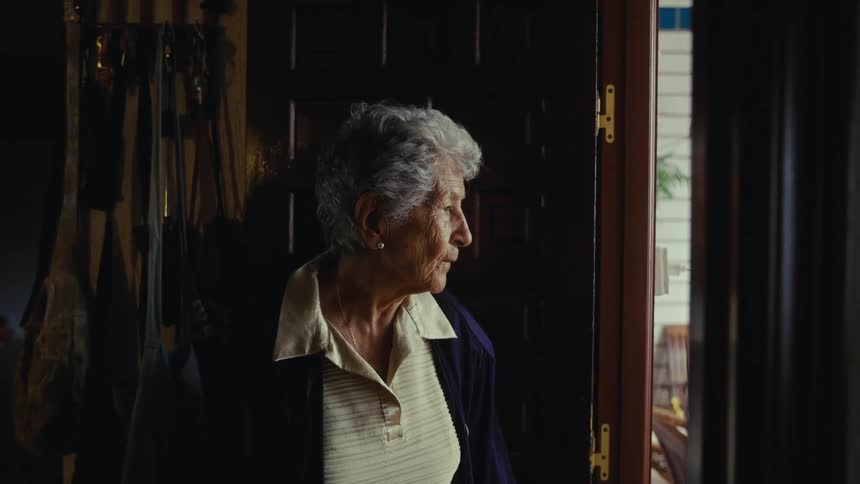 An elderly woman stands pensively in a dark doorway looking outside before slowly walking out of her house