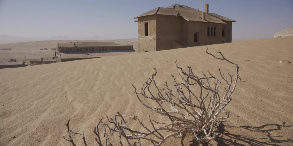 A slow tilt-up reveals a weathered, abandoned building being reclaimed by the vast desert sand dunes
