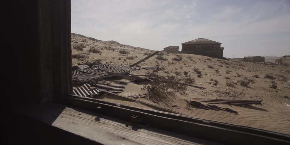 A slow push out of a weathered window reveals the desolate landscape of an abandoned town being reclaimed by the desert sand