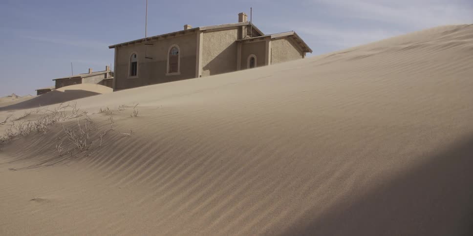 A time-lapse of a shadow moving across a sand dune, gradually revealing an abandoned building being swallowed by the desert