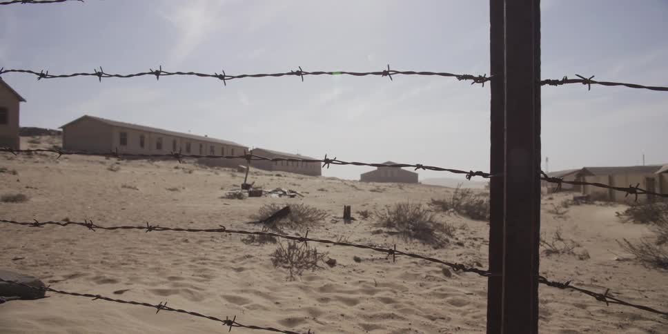 A slow slide past a barbed wire fence reveals the abandoned buildings of a desolate desert ghost town