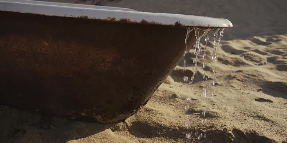 Water spills in slow motion from a rusty old bathtub sitting on a sandy beach