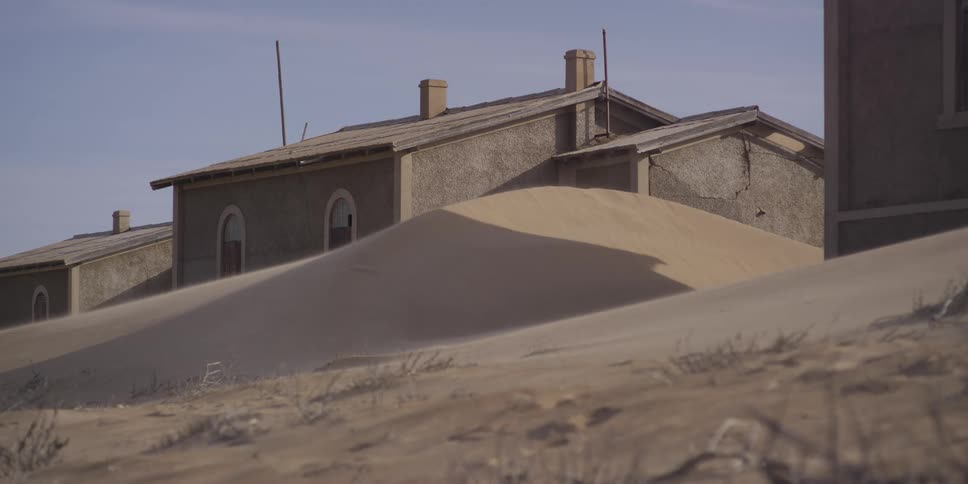 Wind blows sand over large dunes that are slowly engulfing an abandoned building in a desolate ghost town