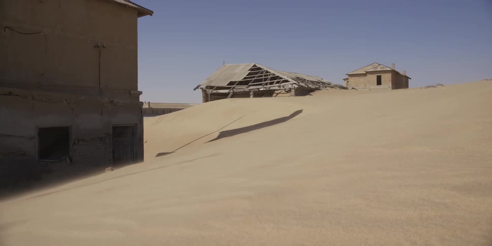 Strong winds sand across the desolate landscape of an abandoned ghost town, with ruined buildings being swallowed by the dunes