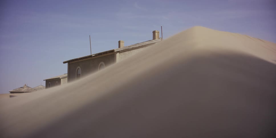 Fine sand blows over the crest of a massive dune, slowly burying an abandoned house in the desert