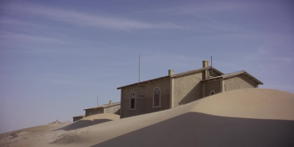 A static shot captures the desolate beauty of Kolmanskop, where abandoned buildings are engulfed by the ever-moving desert sand as the wind blows