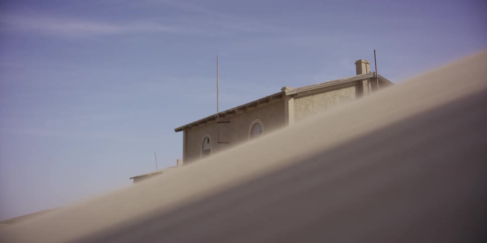A large sand dune obscures an abandoned house in the desert as a strong sandstorm blows