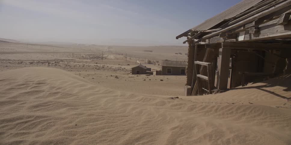 Wind sweeps across sand dunes, slowly burying the decaying buildings of an abandoned desert ghost town